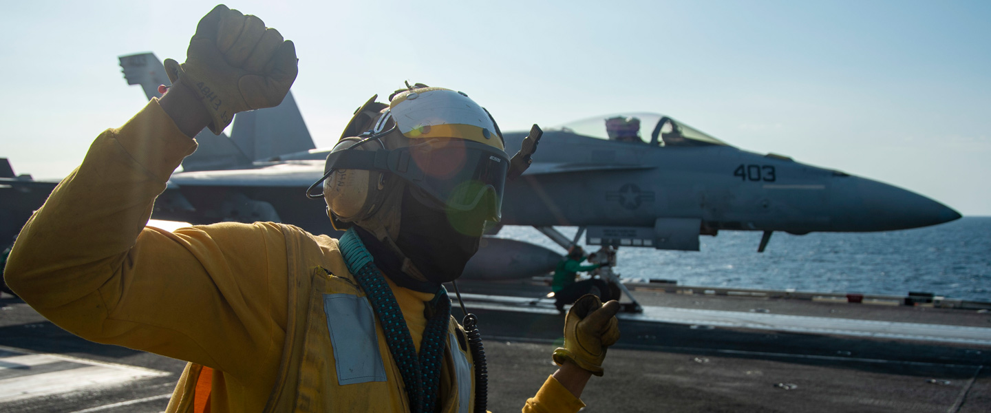 A United States Navy Aircrewman Operator gives hand signals to a Navy Pilot on the runway of an aircraft carrier