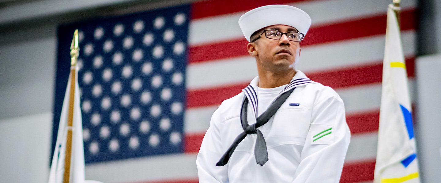 A U.S. Navy Sailor stands at attention in front of the American flag. 