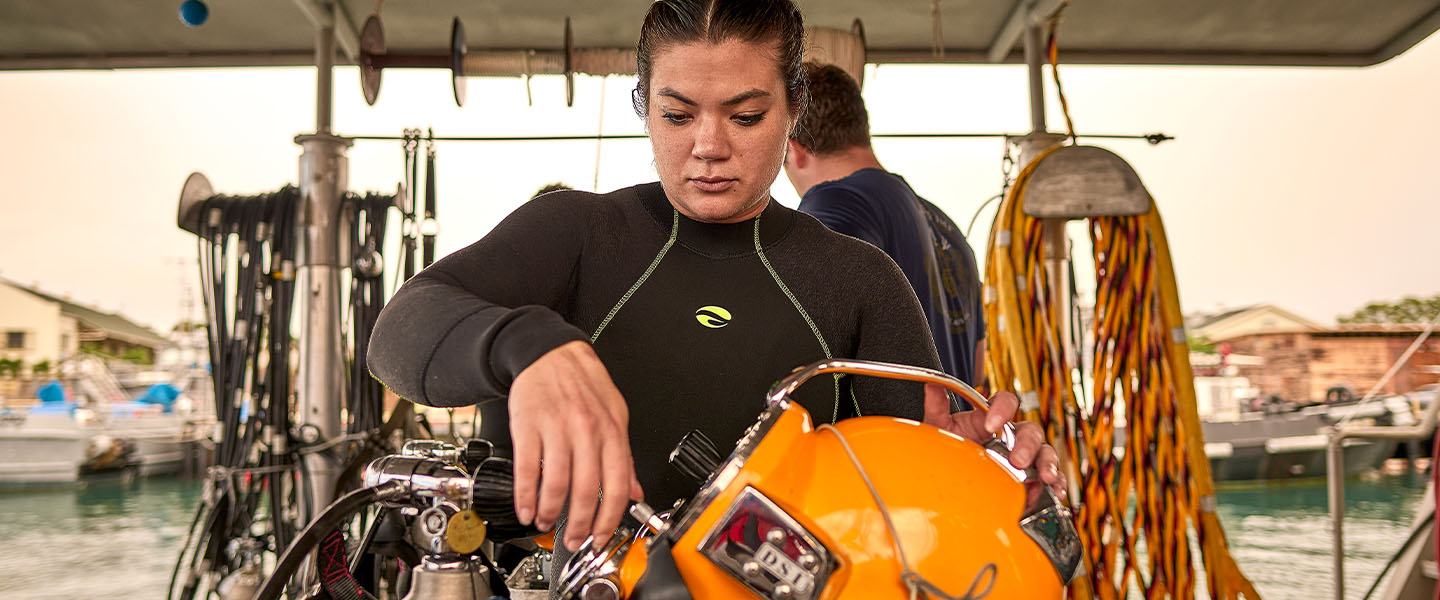 Navy Lieutenant Commander Mia Edgar adjusts a dive helmet while standing on a dock surrounded by marine gear and watercraft.