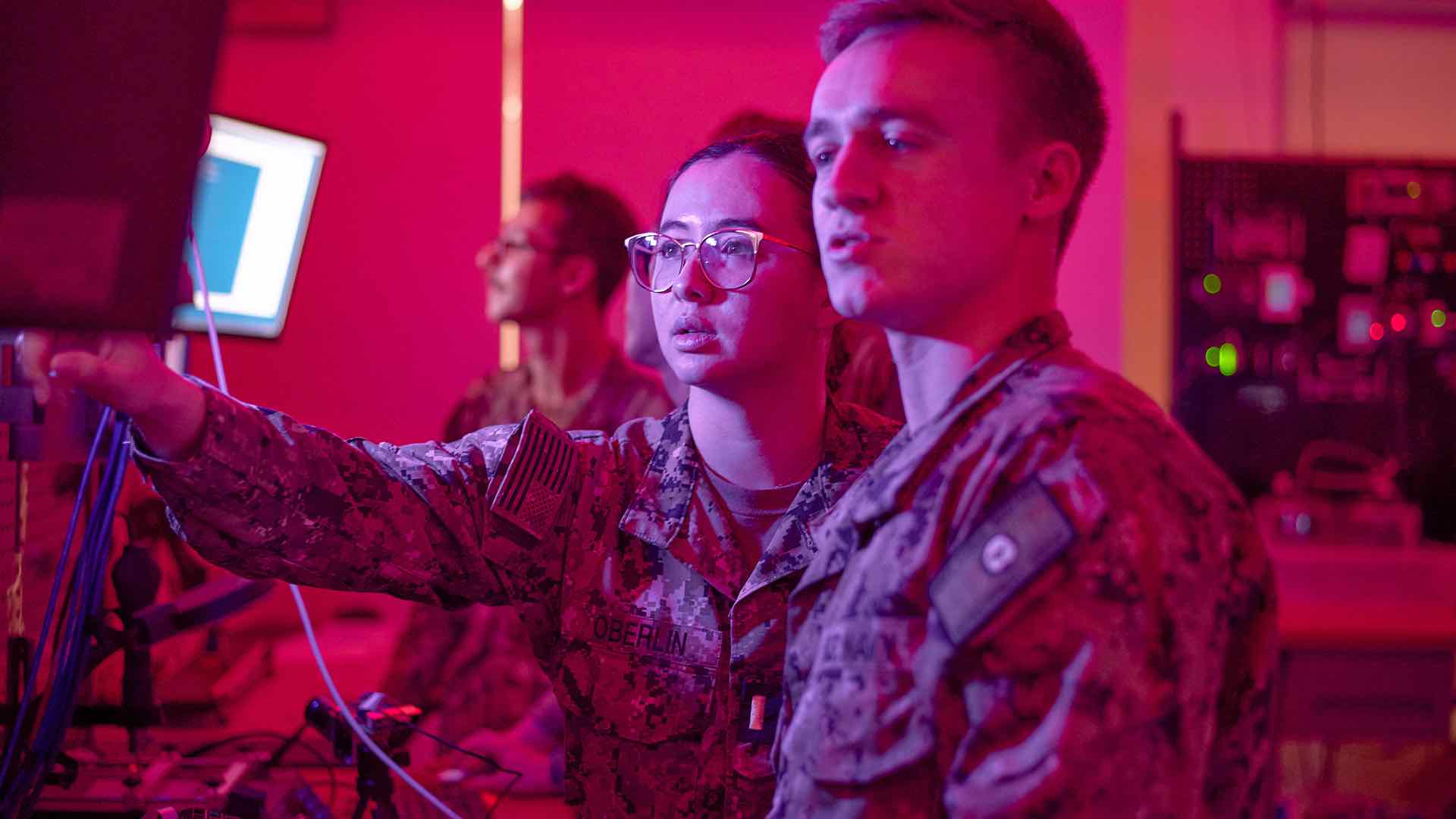 ENS Marie Oberlin stands next to another Sailor and points to a computer monitor.